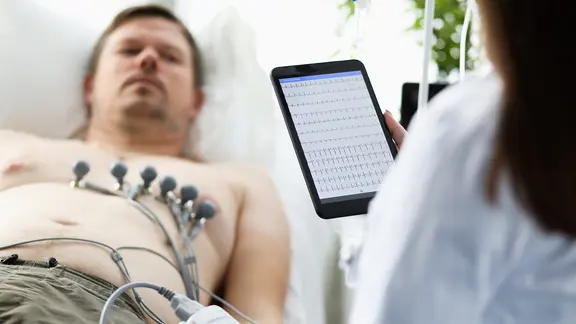 Ein Mann liegt auf einer Liege, eine Ärztin hält ein Tablet mit medizinischen Werten in der Hand.