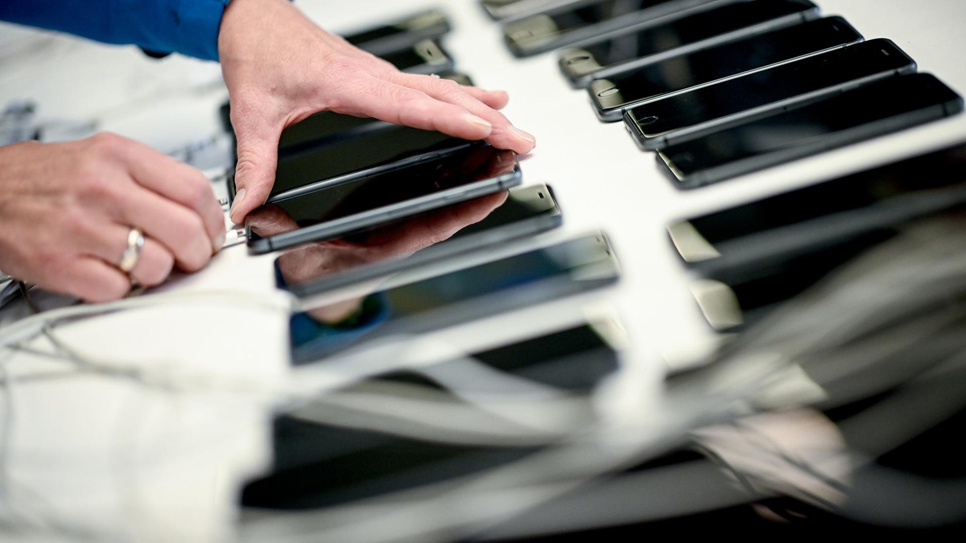 Eine Hand greift nach einem Smartphone, das neben weiteren Smartphones liegt.  | Britta Pedersen/dpa +++ dpa-Bildfunk +++