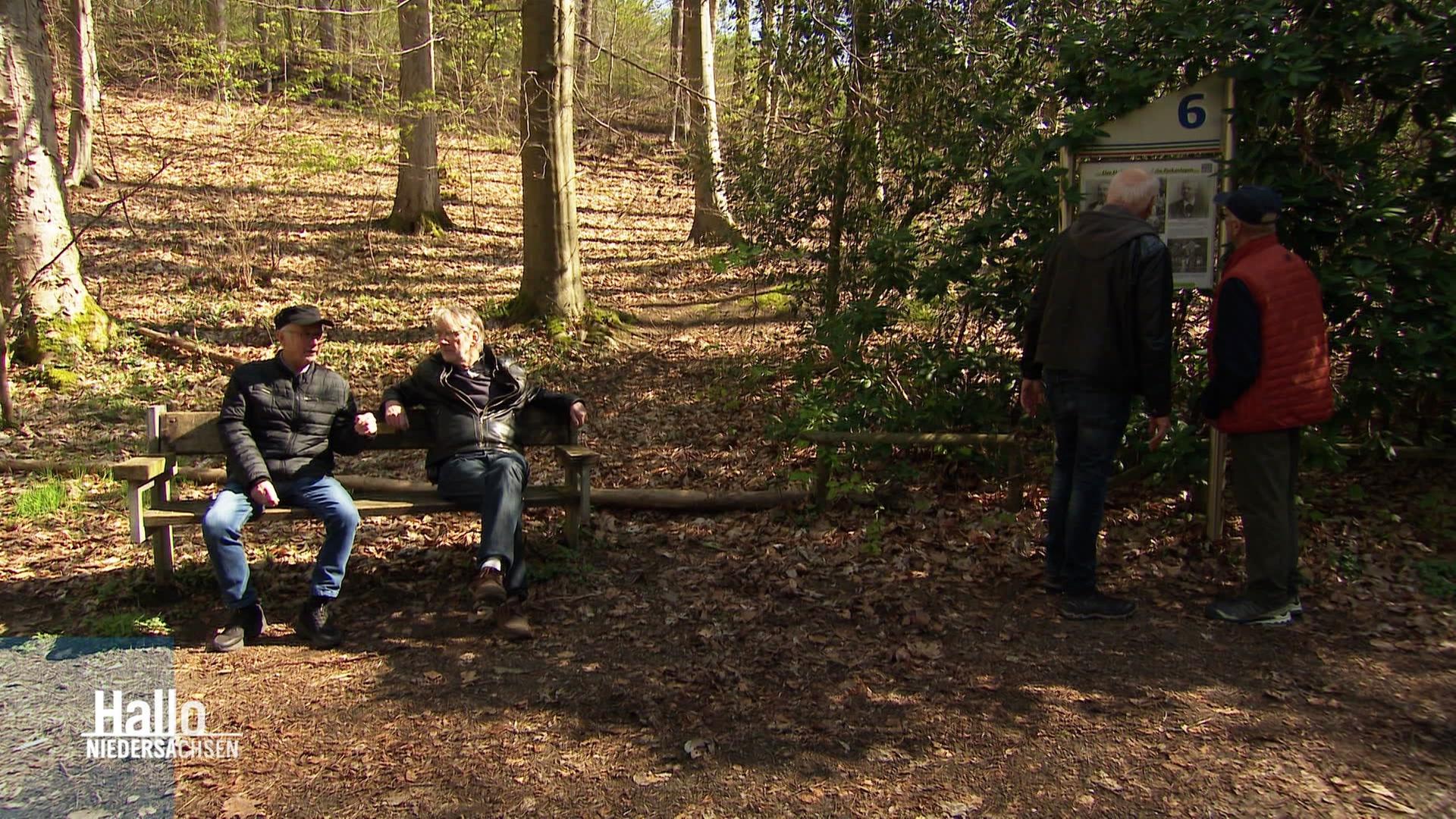 Zwei Männer sitzen im Wald auf einer Bank, daneben stehen zwei Männer vor einer Schautafel. | Screenshot