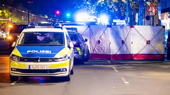 Ermittler haben an der Vahrenwalder Straße einen Sichtschutz errichtet. | dpa/Moritz Frankenberg Polizeiautos parken mit blinkendem Licht auf einer Straße. Im Hintergrund befindet sich ein Sichtschutz.