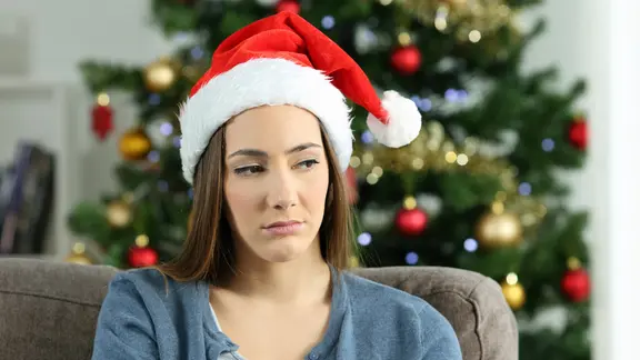 Eine Frau mit Weihnachtsmann-Mütze sitzt traurig auf einem Sofa, im Hintergrund ein geschmückter Weihnachtsbaum