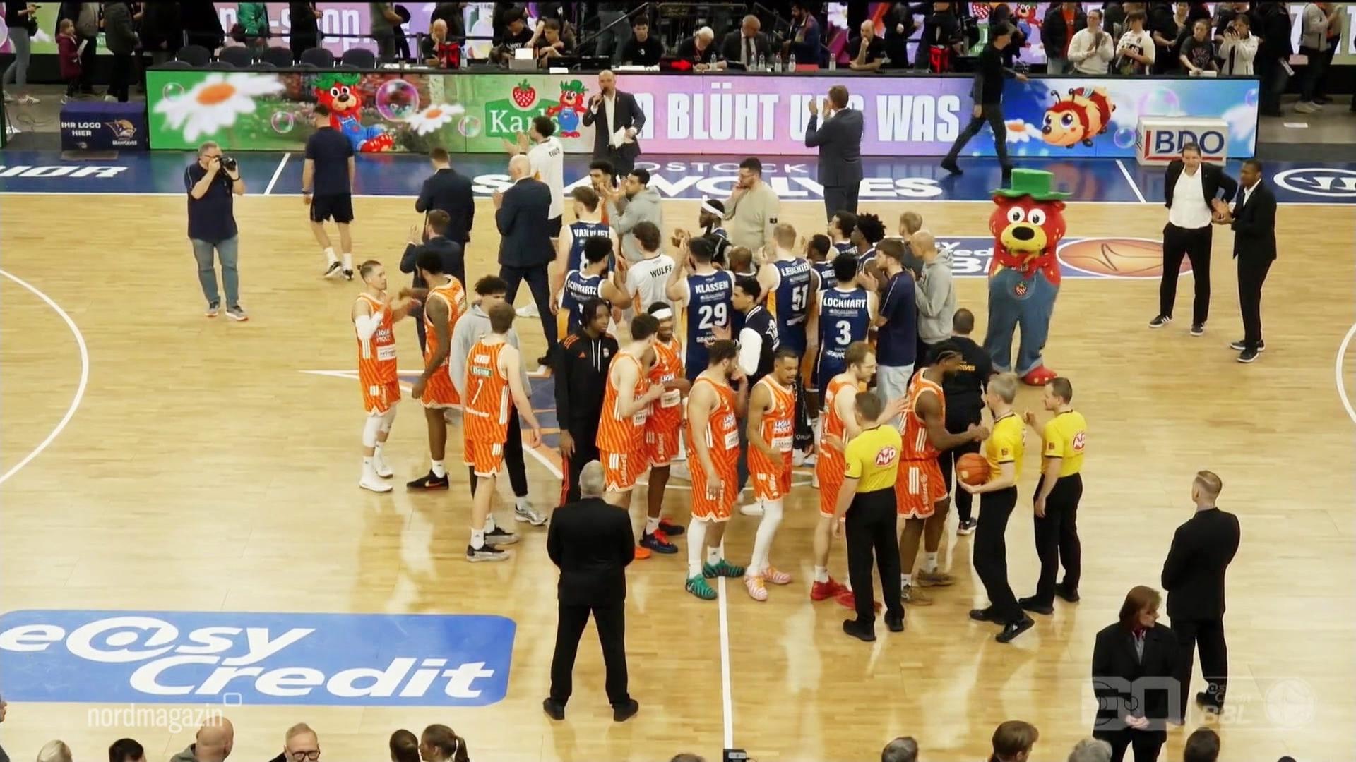 Die Basketballer, Trainer und Teammitglieder aus Rostock und Ulm stehen auf dem Spielfeld. | Screenshot