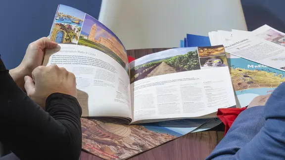 Zwei Personen sitzen an einem Tisch vor aufgeschlagenen Reisekatalogen.