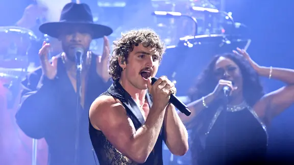 Ein Mann mit Locken, Schnurrbart und nackten Oberarmen singt beim Konzert - US-Musiker Benson Boone bei einem Auftritt