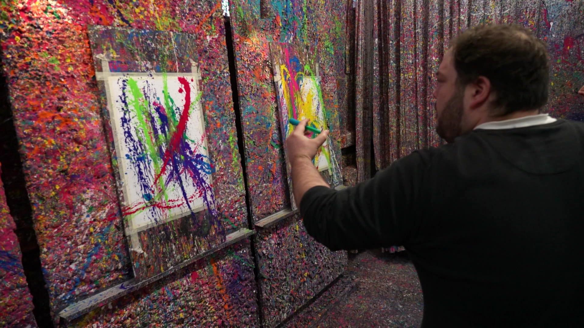 Ein Mann sprüht bunte Farbe auf eine Leinwand, die vor einer Wand steht, die ebenfalls mit vielen Farbspritzern bedeckt ist. | Screenshot