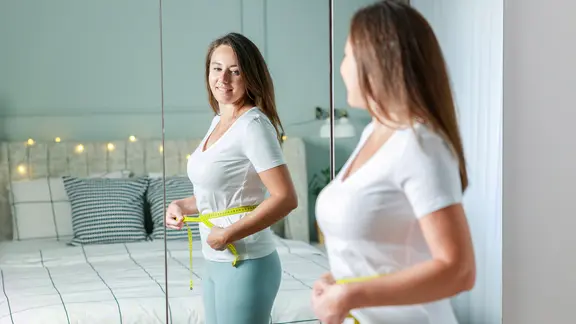 Abnehmen in den Wechseljahren: Eine Frau schaut in den Spiegel und hält ein Maßband um die Hüfte | photoshop.de Abnehmen in den Wechseljahren: Eine Frau schaut in den Spiegel und hält ein Maßband um die Hüfte