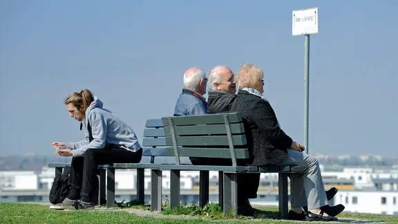 Ein junger Mann sitzt allein auf einer Bank, rechts drei Rentner auf einer Bank, die den Ausblick genießen