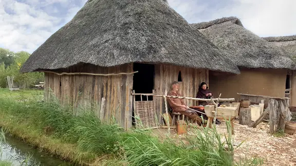 Ein als Wikinger gekleidetes Paar sitzt vor einem rekonstruierten Reetdachhaus im Außenbereich des Wikinger-Museums Haithabu.