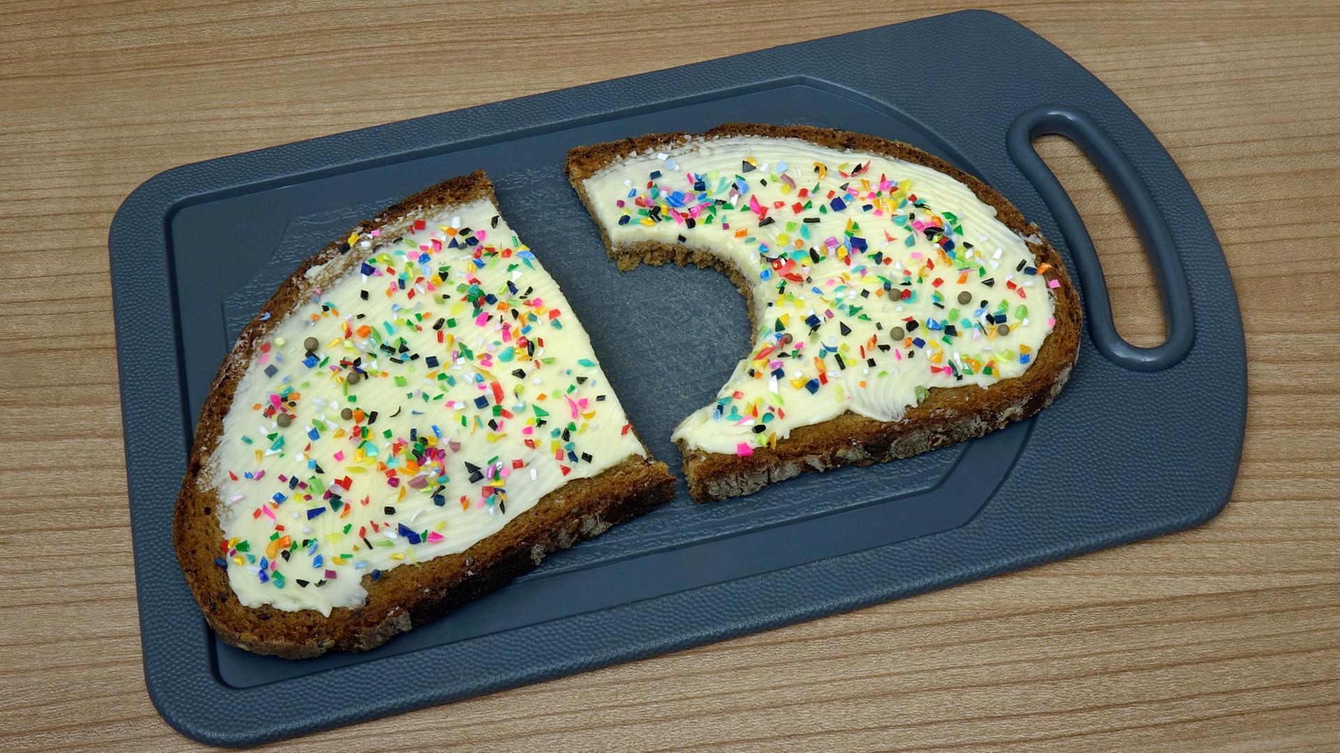 Auf einer angebissenen Scheibe Brot liegen bunte Mikroplastik-Teilchen. (Symbolbild) | picture alliance, ZB | Sascha Steinach