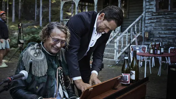 Ein Mann sitzt an einem Klavier und ein anderer steht daneben. Beide spielen zusammen. Das Klavier steht vor einem Haus. 