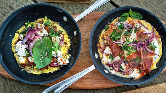 Unterschiedlich belegte Omelett-Pizzen in zwei Pfannen, die auf Holzbrettern stehen.