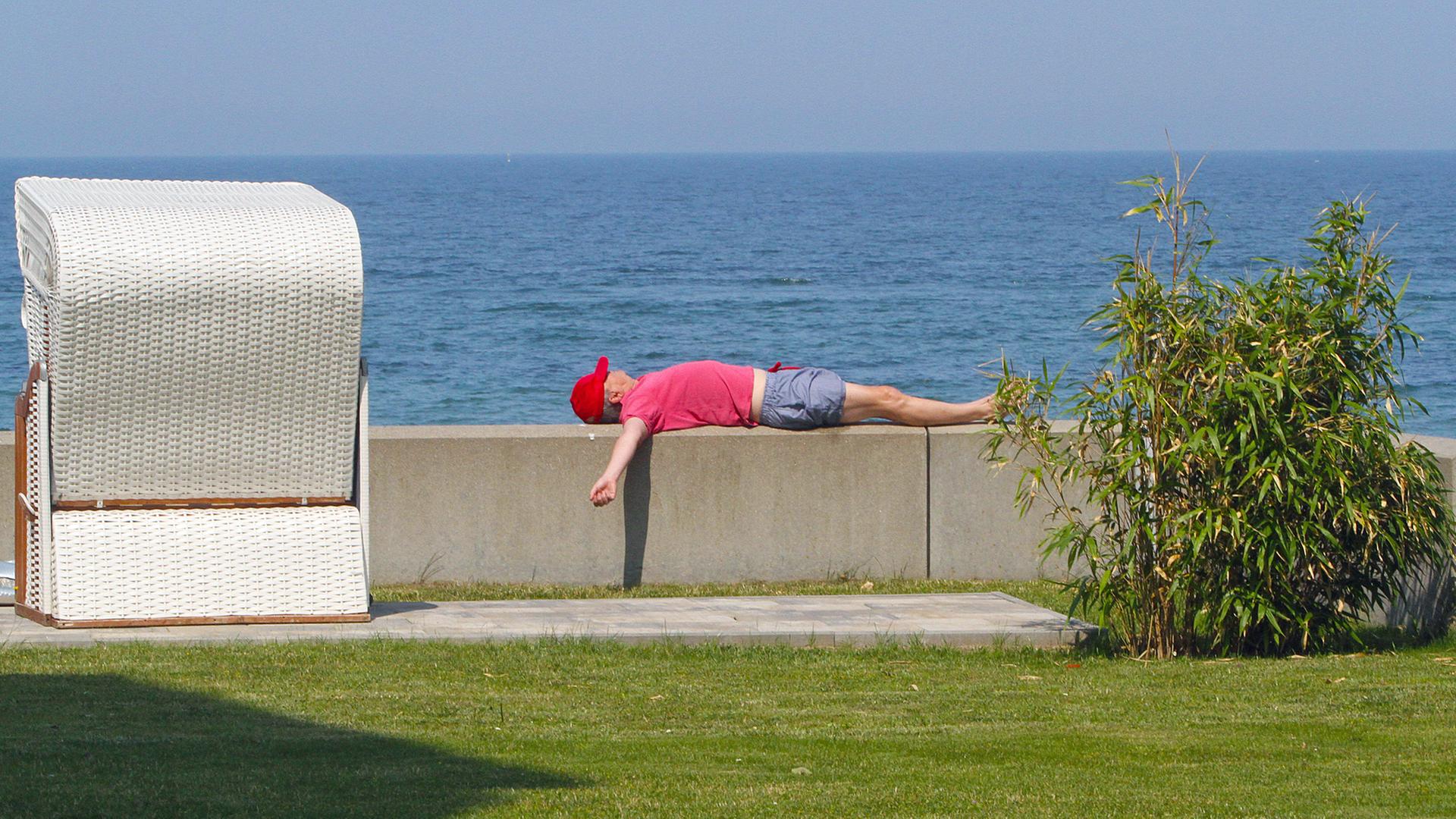 Ein Mann liegt an einem Strandzugang auf einem kleinen Mäuerchen in der Sonne. | IMAGO Images / BildFunkMV