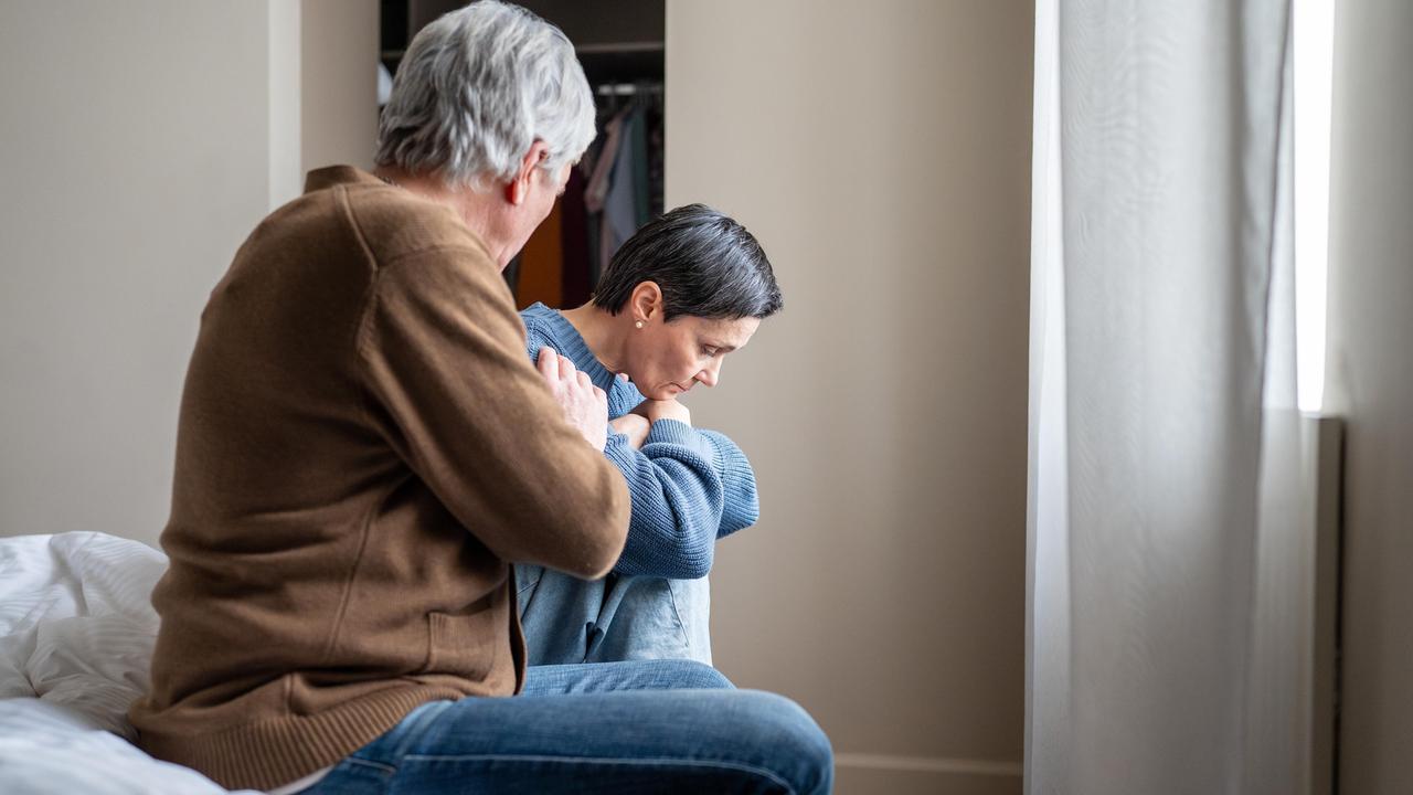 Eine Frau sitzt mit angewinkelten Beinen auf einem Bett. Daneben sitzt ihr Mann.