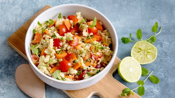 Eine Schale mit Hirsesalat, darin Tomaten, Möhren und frische Kräuter.