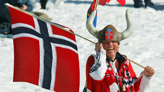 Ένας Νορβηγός με κράνος Βίκινγκ και σημαία γελάει στην κάμερα. | συμμαχία εικόνας/dpa/dpaweb | Roland Holschneider Ένας Νορβηγός με κράνος Βίκινγκ και σημαία γελάει στην κάμερα.