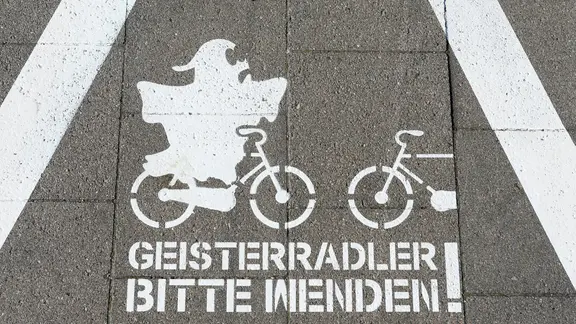 A warning triangle with two oncoming wheels is shown in white on a cycle path. A ghost rides on a bike. The text underneath says: Ghost cyclists, please turn around!