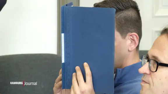 Der Angeklagte verbirgt sein Gesicht hinter einer Mappe.