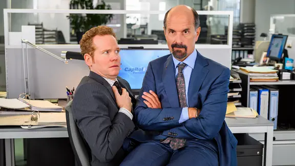 Bernd Stromberg (Christoph Maria Herbst) sitzt in blauem Anzug sitzt auf dem Schoss eines anderen Mannes in grauem Anzug.