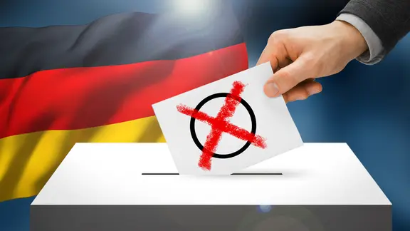 Wahlkarte mit Wahlkreuz wird in eine Wahlurne gesteckt, dahinter eine Deutschlandflagge (Bildmontage)