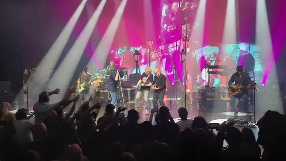 Ein Junge sitzt mit seiner Mutter auf einem Sofa auf der Bühne neben Musikern der Band Fury In The Slaughterhouse. Die Band spielt live, einer der Sänger steht am Mikrofon, ein Gitarrist spielt, im Hintergrund sitzt ein Keyboarder. Die Bühne ist in blau-violettes Licht getaucht.