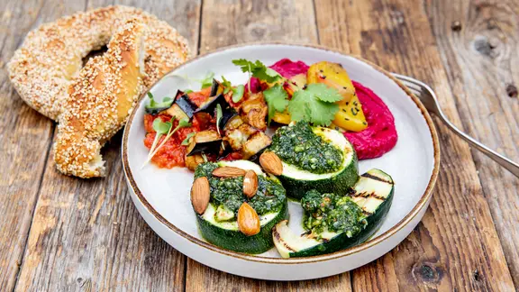 Ein Teller mit Rote-Rete-Hummus, Auberginen in Tomatensoße und Zucchini mit Pesto. Daneben liegt ein Sesamkringel
