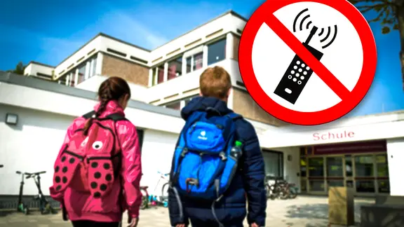 Eine junge Schülerin und ein junger Schüler gehen Richtung eines Schulgebäudes, daneben ist ein Symbolbild mit einem roten Schild zu erkennen, auf dem ein durchgestrichenes Handy abgebildet ist