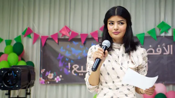 Eine junge Frau mit dunklen Haaren steht auf einer Bühne und hält ein Mikrophon in der Hand.