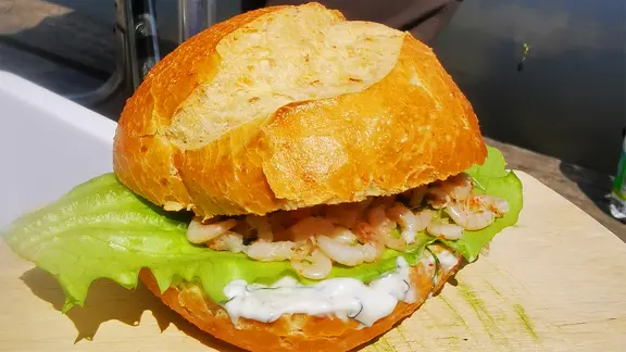 Ein weißes Brötchen mit Krabben, grünem Salatblatt und heller Soße auf einem Holzbrett