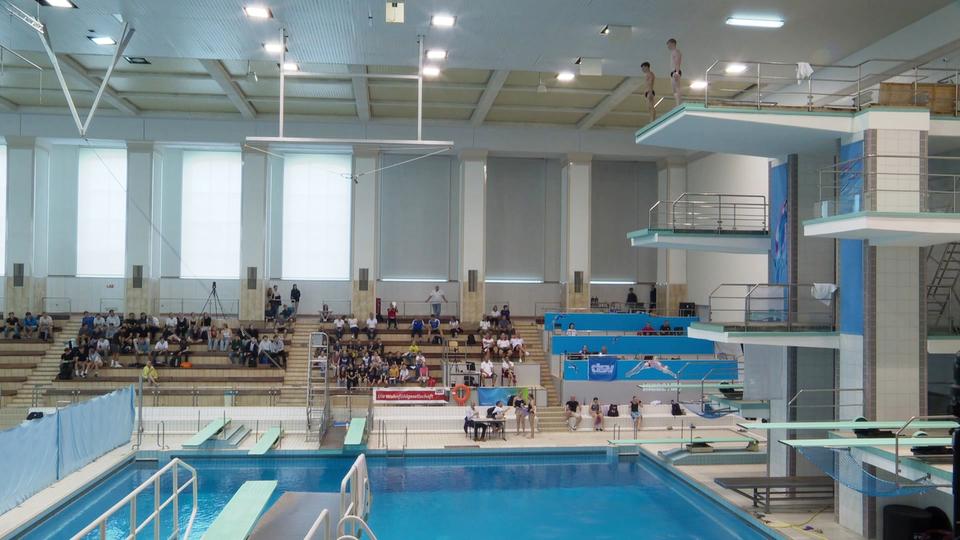 Regelmäßig finden in der Rostocker Neptunschwimmhalle auch nationale und internationale Wettkämpfe statt. Bald ist das Becken leer. | Screenshot Innenansicht der Neptun-Schwimmhallein Rostock mit Tribüne und Sprungtürmen
