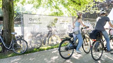 Zwei Menschen fahren Fahrrad. Im Hintergrund das Banner für die Velostation der Kieler Woche.