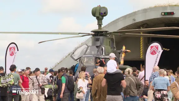 Blick über eine Menschenansammlung, die sich auf einem Militärflugplatz befinden. In ihrer Mitte steht ein Bundeswehr-Helicopter.