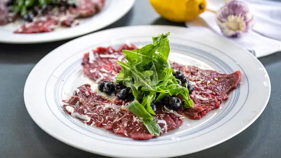 Carpaccio mit Heidelbeeren, Parmesan und Rucola auf einem Teller angerichtet.
