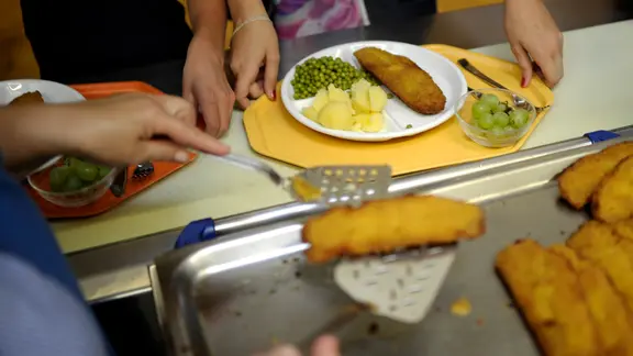 Eine Küchenhilfe legt ein Stück panierten Fisch auf einen Teller eines Schulkindes in einer Schulküche.