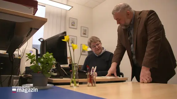 Ein Mann stützt sich auf einen Schreibtisch, neben ihm sitzt eine Frau.