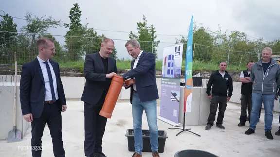 Drei Männer in Anzügen stehen nebeneinander und reichen sich ein orange farbiges Rohr.