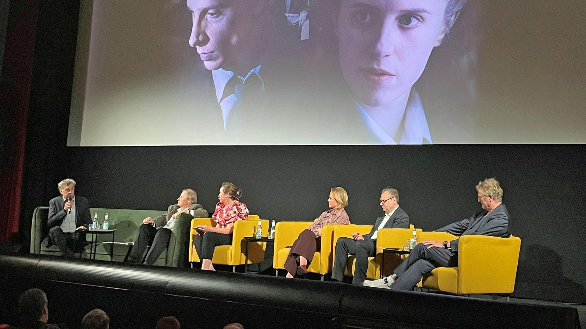Sechs Menschen sitzen in gelben Sesseln auf einer Bühne vor einer großen Filmleinwand. | NDR.de / Jens Büchsenmann