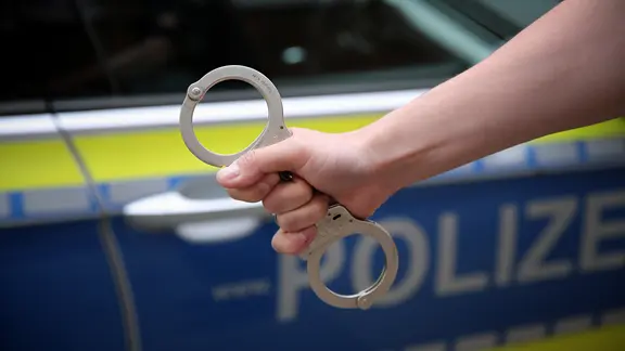 Ein Polizist hält Handschellen einsatzbereit vor einem Streifenwagen.