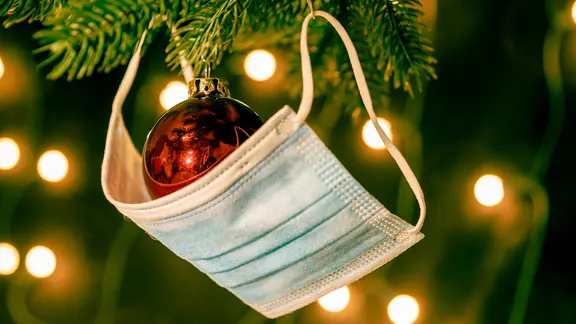 Eine Mund-Nasen-Bedeckung hängt über einer Christbaumkugel am Weihnachtsbaum.