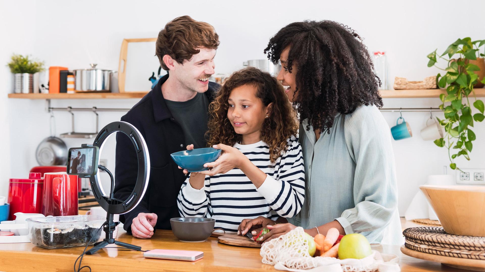 Eine Familie steht in einer Küche vor einem aufgestellten Smartphone. | picture alliance / Westend61 | NOVELLIMAGE