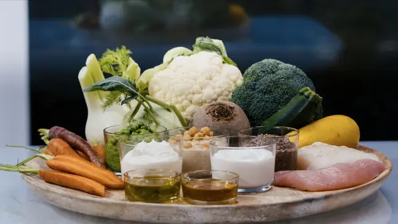 Verschiedene gesunde Lebensmittel wie Gemüse, Joghurt und Öle stehen oder liegen auf einer Holzplatte.