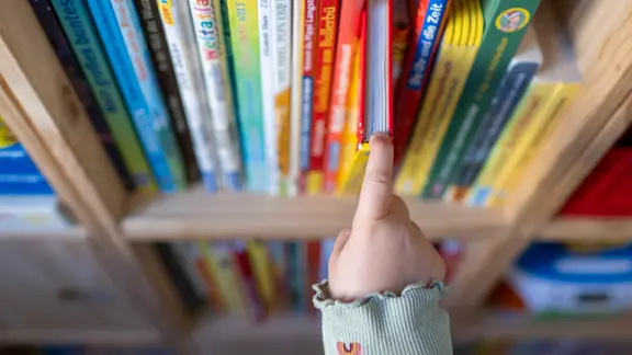 Eine Kinderhand zieht ein Buch aus einem Bücherregal. | picture alliance/dpa | Hendrik Schmidt Eine Kinderhand zieht ein Buch aus einem Bücherregal.