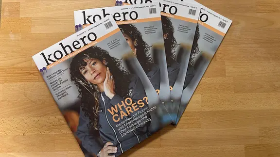 Vier Exemplarte der gleichen Ausagbe des Magazins kohero liegen auf einem Holztisch. Auf dem Cover steht in großen Buchstaben „WHO CARES?“. Das Titelthema der Ausgabe beschäftigt sich mit Care-Arbeit, Gleichberechtigung und Fürsorge. Auf dem Titel ist eine Frau mit lockigen Haaren abgebildet.