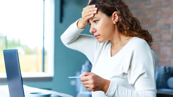 Eine Frau sitzt mit besorgtem Blick vor einem Laptop, in der Hand eine Kreditkarte. 