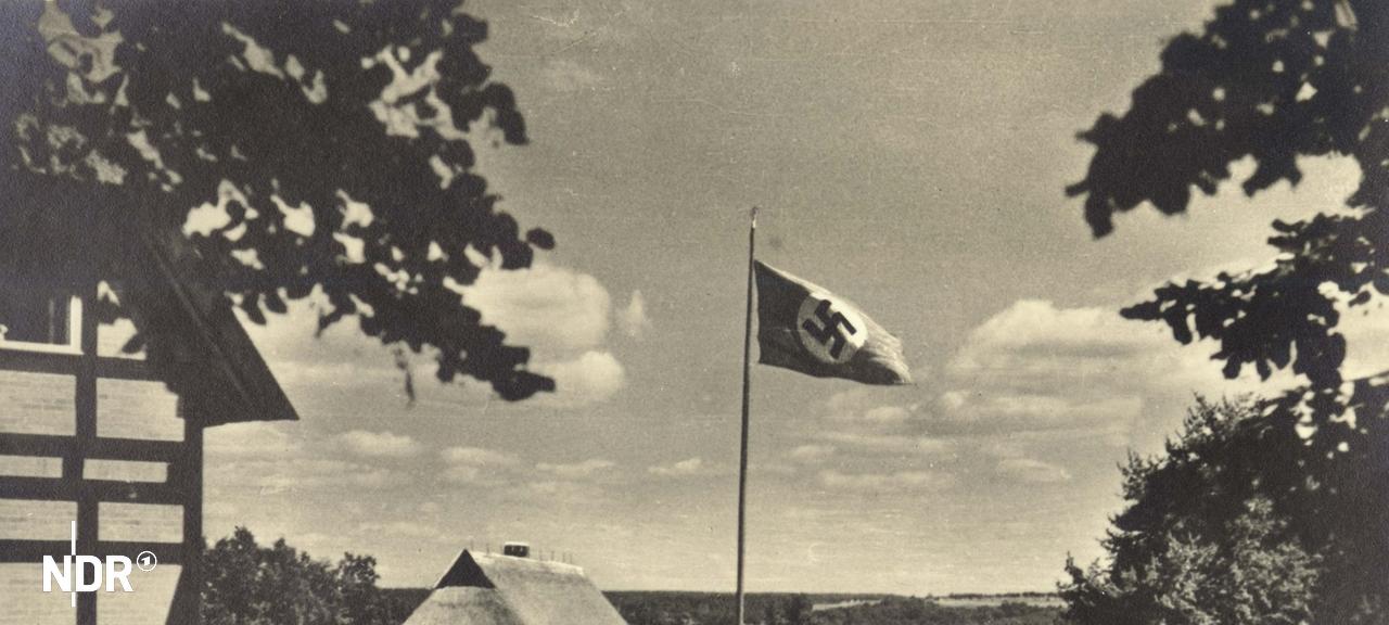 Eine historische Ansichtskarte von Alt Rehse. Auf einem Appellplatz steht ein Fahnenmast mit einer Hakenkreuzfahne. | Archiv EBB Alt Rehse e.V., Archiv EBB Alt Rehse e.V. Eine historische Ansichtskarte von Alt Rehse. Auf einem Appellplatz steht ein Fahnenmast mit einer Hakenkreuzfahne.