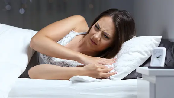 Junge Frau im Bett schaut wütend auf den Wecker, der auf dem Nachttisch steht.