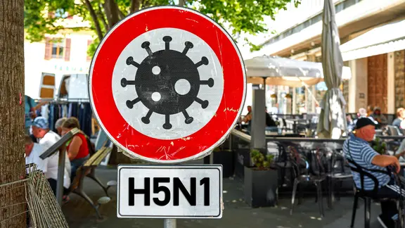 Vor einem Café sitzen Menschen, davor steht ein Warnschild mit einem Virussymbol und der Aufschrift "H5N1". (Symbolbild)