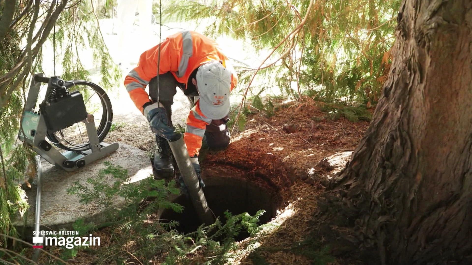 Ein Mann im orangenen Anzug steht an einem Baum vor einem Loch und hält ein Rohr in der Hand. | Screenshot