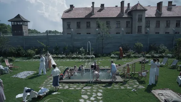 Eine große Familie springt auf der grünen Wiese um einen kleinen Pool herum - dahinter ist ein KZ zu erkennen. Szene aus dem britisch-polnischen Spielfilm "The Zone of Interest" von Jonathan Glazer