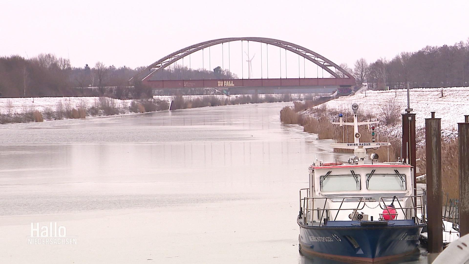 Ein Fluss mit einem Schiff und einer Brücke, links und rechts Schnee. | Screenshot