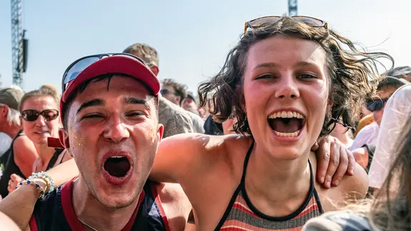 Zwei junge Festivalbesucher*innen stehen lachend und jubelnd in der Menge beim Hurricane Festival. Sie haben die Arme umeinander gelegt und strahlen in die Kamera. Im Hintergrund sind weitere Menschen und blauer Himmel zu sehen.